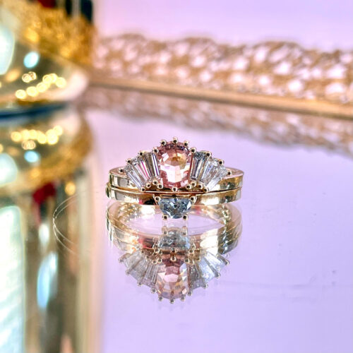 This incredible one of a kind commitment set features a dazzling peachy pink sapphire, surrounded by fancy diamond cuts in half-moon and baguettes.  All lovingly set in 14K yellow gold, this custom set delighted our clients. 